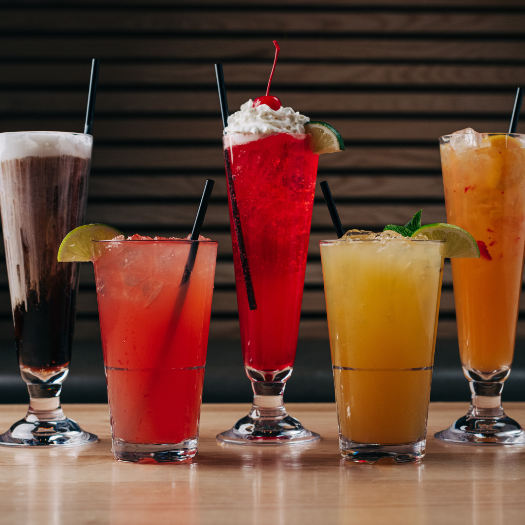Five colorful cocktails and frozen drinks lined up on the bar with lime and mint garnishes